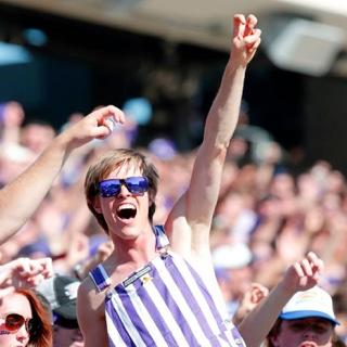 A happy, cheering student in striped overall and purple sunglasses makes the two-fingered "Go Frogs" hand sign at a crowded football game.