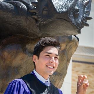 A male TCU student in Honors regalia makes the two-fingered "Go Frogs" hand sign in front of a bronze statue