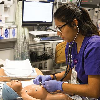 A nursing student in purple scrubs uses a stethoscope as she practices on an infant simulation dummy