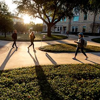 TCU students wearing jackets walk on campus, casting long shadows on a sunny day