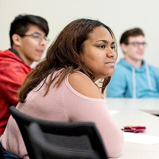 Three TCU students of different ethnicities listen together in a classroom. A young woman in a pink sweater is in the foreground.