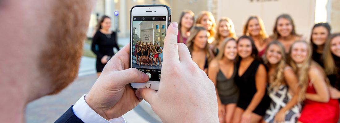 A young man uses a cell phone to photograph a group of smiling sorority girls before a party in the Greek Village at TCU.